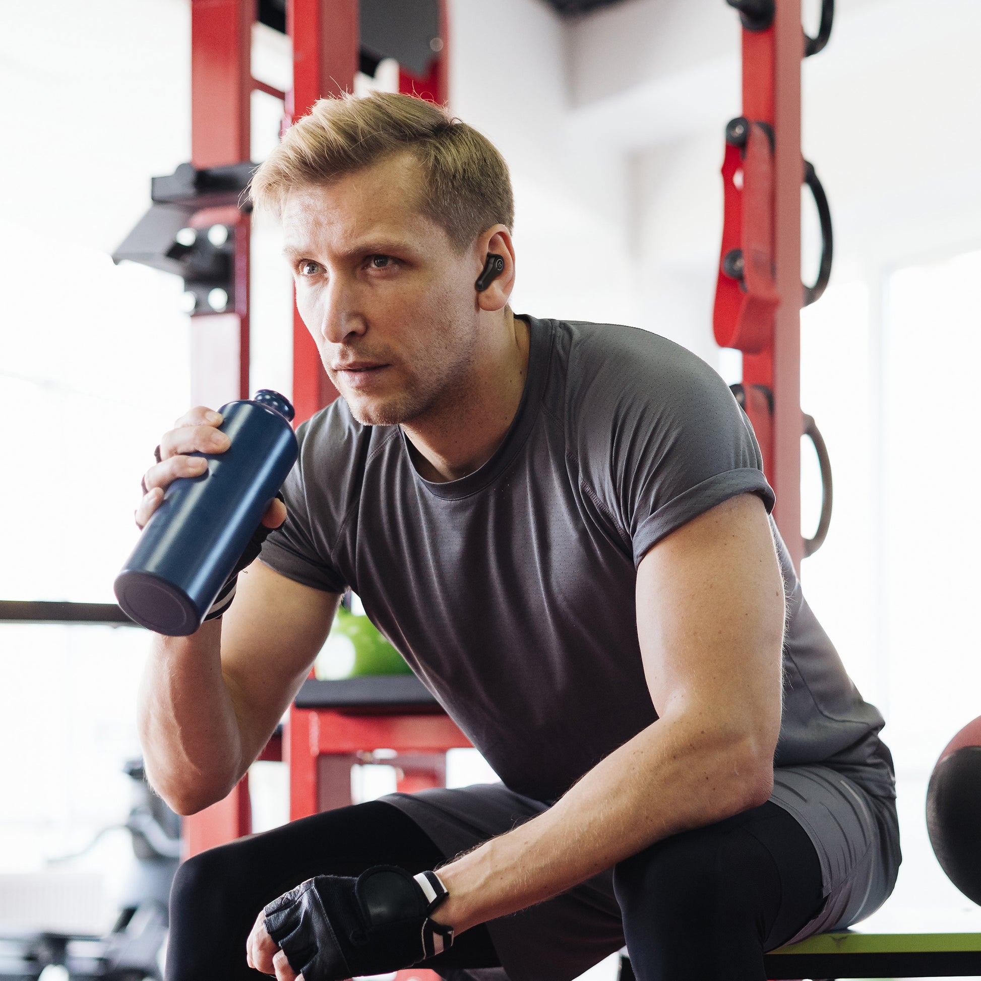 Photo of Morpheus 360 Pulse HD True Wireless Earbuds, shows a young white male at the gym with his black Pulse HD Earbuds in and drinking water between workouts.