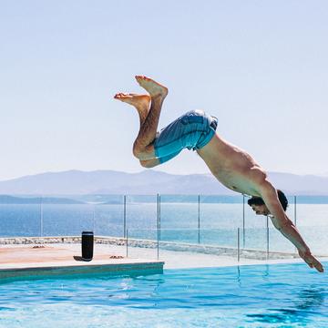 Picture of a young man in swim trunks diving into the pool with the Morpheus 360 Sound Ring II Speaker behind him playing music.